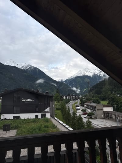 The image depicts a view from a wooden balcony overlooking a town nestled in a valley, with snow-capped mountains in the background. The scene is set in Mayrhofen, Austria, as indicated by the "Neuhaus Zillertal Resort" signage on a dark building in the foreground.

In the immediate foreground, the dark wooden railing of the balcony is visible. Beyond the railing, a dark-colored building with white lettering that reads "Neuhaus" and "ZILLERTAL RESORT" dominates the left side of the frame. This building appears to be part of the resort. To the right of this building, a winding road can be seen with several cars parked alongside it and a few in motion. Further into the background, other buildings, including what appears to be a train station or a bus depot, are visible.

The environment is mountainous, with steep, densely forested slopes rising behind the town. The peaks of these mountains are covered in snow, suggesting either a high altitude or recent snowfall. The sky is overcast with clouds, casting a diffused light on the scene. The overall impression is one of a serene mountain town, possibly during the shoulder season or a cooler time of year. There are no people clearly visible in the image. The setting appears to be a resort area, characteristic of the Austrian Alps.