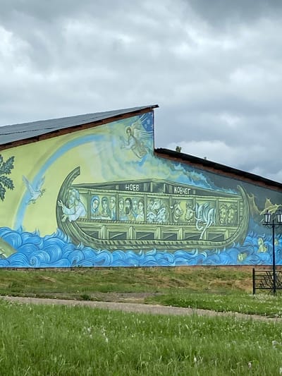 This is a photograph of a mural on the side of a building, depicting a scene from the biblical story of Noah's Ark. The mural, located in Кварса, Russia, shows a large ark with multiple levels filled with various animals, including what appear to be lions, elephants, and other creatures. Several people are visible inside the ark, looking out from different windows. An angelic figure is depicted in the upper left, and a dove carrying an olive branch flies in the lower part of the mural, symbolizing peace. The ark is surrounded by stylized blue waves. The mural is painted on a building that appears to be somewhat aged. To the right of the mural is a grassy area with a path. The sky above is cloudy, suggesting overcast weather. Text visible on the ark reads "КОВЧЕГ," which translates to "Ark" in Russian, and "НОЕВ," which translates to "Noah's."