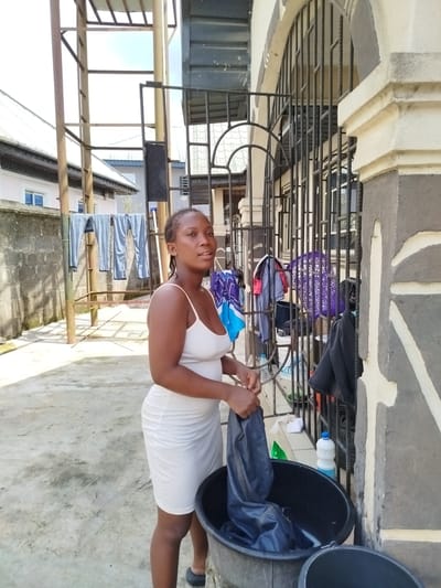 A young woman stands in a courtyard, holding a pair of blue jeans over a large black basin. She is wearing a white, spaghetti-strap dress. The setting appears to be a residential area in Sapele, Nigeria, indicated by the architecture and the laundry drying on a rack in the background. The time of day seems to be daytime, with bright sunlight casting shadows on the concrete ground.
In the background, several pairs of pants are hanging on a metal clothesline. There are buildings with tiled roofs and barred windows. A metal grate with decorative bars covers a window or entryway on the right side of the frame. Various items of clothing are hanging on this grate, including a purple woven basket. A white bottle with a blue label and a green plastic bag are also visible near the basin.
The woman is looking away from the camera with a neutral expression. The activity taking place is likely laundry, with the woman preparing to wash the jeans in the basin, which contains water. The overall impression is one of domestic chores being performed outdoors. There is no visible text in the image.