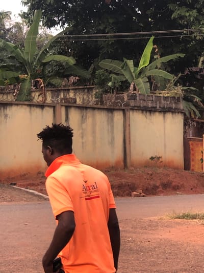 The scene depicts an outdoor environment in Neni, Nigeria. In the foreground, an adult male, facing away from the camera and slightly to the left, is visible. He has dark skin, short dark textured hair with faded sides, and is wearing a short-sleeved, bright orange polo shirt with a collar. The back of his shirt displays a logo with the text "Airal" and "water pump" below it, separated by a red horizontal line. A dark object, possibly a mobile phone, is held in his left hand near his hip.
Behind the individual, a low, light beige concrete or stucco wall with visible vertical dark stains extends horizontally across the mid-ground. Sections of the wall are topped with barbed or razor wire. Behind and above this wall, dense green foliage is visible, including several large-leafed plants consistent with banana or plantain trees. Further back, utility lines are strung across the upper portion of the frame. The ground in the foreground and immediately adjacent to the wall consists of reddish-brown soil, with a grey paved road surface partially visible in the mid-ground, sloping slightly. Scattered reddish debris, possibly bricks, are present on the soil near the wall.