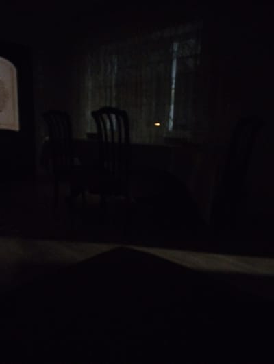 The image depicts a dimly lit indoor scene, likely a dining area. In the background, a window with visible blinds is present, with a faint light source behind it. In the mid-ground, a table is visible, with several chairs around it. The chairs appear to be dark, possibly wooden. On the far left, a portion of a framed object is visible. The foreground is dark and indistinct. The scene is devoid of any people. The overall lighting suggests it is nighttime or a very dark room. There is no visual information in the image to suggest a specific location in Znamenskoye, Russia.