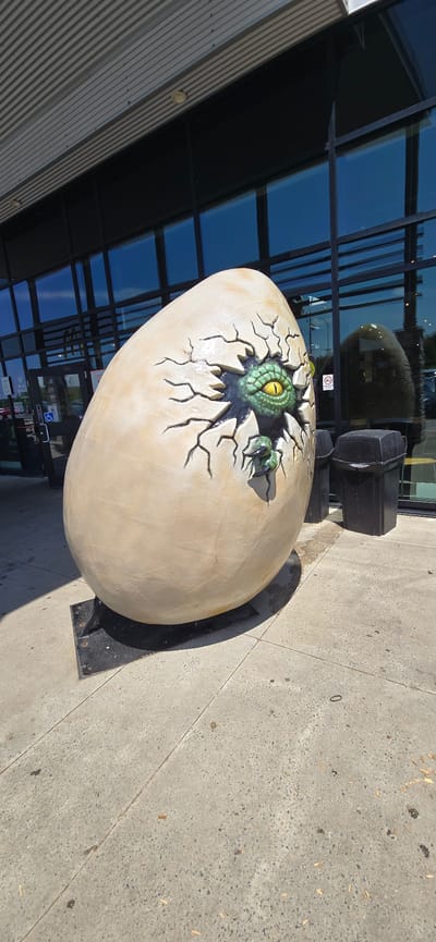 A large, light-colored egg sculpture with a dinosaur hatching out of it is on display outdoors. The dinosaur's green head with a yellow eye and two claws are visible emerging from a crack in the egg. The sculpture is positioned on a black metal base. In the background, a modern building with large reflective blue windows and a ribbed silver facade is visible. A black trash receptacle is situated behind the egg. The ground is a concrete sidewalk with scattered leaves. The scene appears to be outdoors during the daytime, with sunlight casting shadows. The setting is likely a public space or business entrance. No people are visible in the frame. There are some reflections in the windows and a faint "0" etched into the concrete.