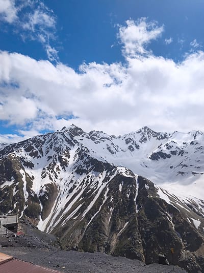 The image depicts a mountainous landscape in the vicinity of Terskol, Russia, under a partly cloudy blue sky. In the foreground, there is a rocky, dark gray ground with some scattered debris and a section of a corrugated metal roof. Further back, a concrete structure, possibly a small building or platform, is visible on the left side of the frame.

The dominant feature of the image is a large, snow-capped mountain range. The mountains are rugged, with dark rocky slopes interspersed with patches of white snow. The snow cover is uneven, suggesting the effects of melting or wind erosion, creating a dramatic contrast between the dark rock and the bright snow. Streaks of snow run down the mountainsides in various directions. The peaks are sharp and jagged, indicative of a high-altitude environment.

In the sky, scattered white clouds are present, with areas of clear blue sky visible between them. The lighting suggests it is daytime, with the sun illuminating the scene. There are no people or animals visible in the image. The overall scene conveys a sense of natural grandeur and the harsh beauty of a high mountain environment.