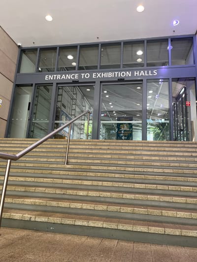 The image displays a modern entrance to an exhibition facility, featuring a wide outdoor staircase leading up to glass doors. The staircase is constructed from alternating light and dark-toned stone or concrete treads, with a contemporary metal handrail running alongside it on the right.

Above the main glass entry doors, a clear sign with white capital letters on a dark grey background reads, "ENTRANCE TO EXHIBITION HALLS." The transparent doors offer a glimpse into the building's interior, revealing a spacious hall with a high ceiling, numerous recessed lights, and visible structural elements. Through the glass, a colourful poster or sign advertising an exhibit is discernible, showing cartoon-like animals and partial text, including "MOON" and "ENTRY," accompanied by a directional arrow pointing left. A small red and white safety sign is also faintly visible inside.

No people are present, indicating a quiet moment. The scene appears to be set during the daytime. This functional and inviting entrance is located in South Brisbane, Australia.