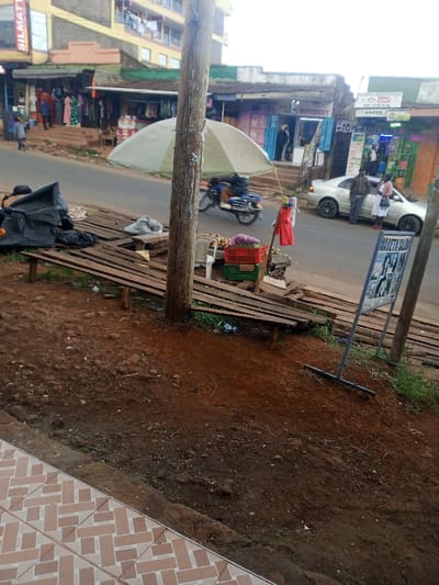 A street scene features a roadside fruit stand shaded by an umbrella, next to wooden planks and a sign advertising a salon. A motorcycle passes in the street, where a silver car is parked with two people standing nearby. Shops line the street in the background under a cloudy sky. The foreground shows bare earth transitioning to a tiled walkway.