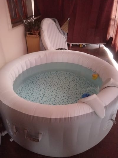An inflatable hot tub filled with water is the main subject, set against a backdrop of a room with a window and curtains. The hot tub is round and grey with a white inflatable rim. The water has a light blue speckled pattern at the bottom. A yellow rubber duck and a blue object, possibly a filter or toy, are floating in the water. The scene appears to be indoors, possibly on a patio or in a room with a floor. The lighting suggests it could be daytime, with sunlight casting shadows. To the left, a window with wooden frames is visible, and behind the hot tub, dark red curtains hang. A potted plant in a wooden crate sits near the window. There is no visible text in the image. The overall impression is a relaxed, possibly domestic setting.
