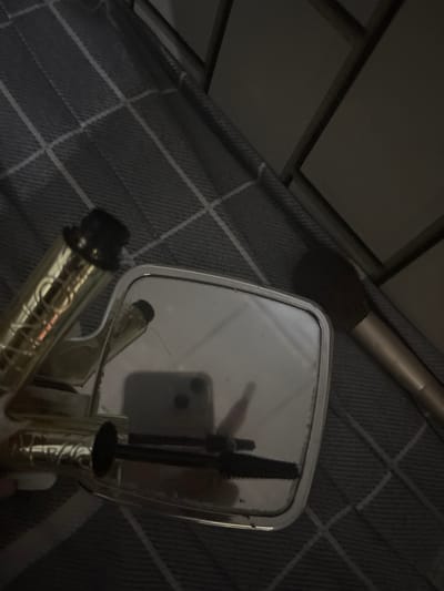 This is a close-up, overhead shot of a hand-held mirror and a mascara wand, set against a textured, plaid-patterned surface. The lighting is dim, suggesting it might be evening or a dimly lit room. The mirror reflects a portion of a smartphone, identifiable by its camera lenses. The mascara wand, with its black bristles and golden tube, is positioned over the mirror. The overall scene is focused on these cosmetic items. There is no visible text in the image. The location context of Velykyi Sukhodil, Ukraine, is provided but not evident from the visual information alone.