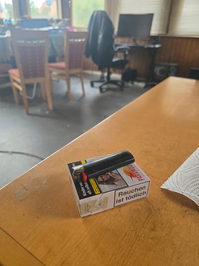 This is a horizontal, slightly blurry, first-person perspective photograph taken indoors, likely in a room with a wooden table. In the foreground, resting on the table, is a pack of "PARAMOUN QUALITY AMERICAN BLEND" cigarettes and a black lighter. The cigarette pack features a red and orange sun logo and a warning label in German: "Rauchen ist tödlich" (Smoking is deadly), and another warning: "Kinder von R oft selbst z" (Children from R often themselves).
The background of the image is out of focus, but it appears to be a living or dining area. There are chairs with red upholstered seats and wooden frames visible. A window with vertical blinds lets in natural light, suggesting it might be daytime. In the further background, some office equipment like a computer monitor and a desk are vaguely discernible, along with what looks like wooden paneling on the wall. A white paper towel is also visible on the table, partially obscuring the top right corner of the cigarette pack. The overall impression is of a casual, domestic setting where smoking may be present. The image is oriented vertically, with the top of the scene appearing on the left.