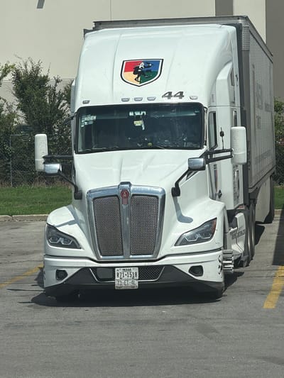 A white semi-truck with a silver grille is parked on asphalt. The truck has a red Kenworth logo on the grille and a colorful shield emblem on the hood with the number 44 below it. A license plate on the front of the truck reads "TEXAS R71-1518 73-ET-4L". A large metal trailer is attached to the truck. The truck is parked in front of a light-colored building, and there are green trees and a chain-link fence behind it. The scene appears to be in an industrial or commercial area, possibly a parking lot. The location is identified as Lebanon, United States of America.
