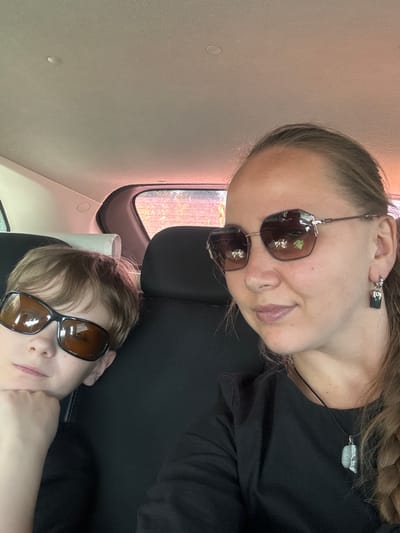 Inside a vehicle in Средний Постол, Russia, a woman and a child are visible. The woman, wearing sunglasses, a dark shirt, and a necklace, is positioned at the top of the frame. A child with sunglasses rests on the headrest below her. The interior of the vehicle is visible, including a window.