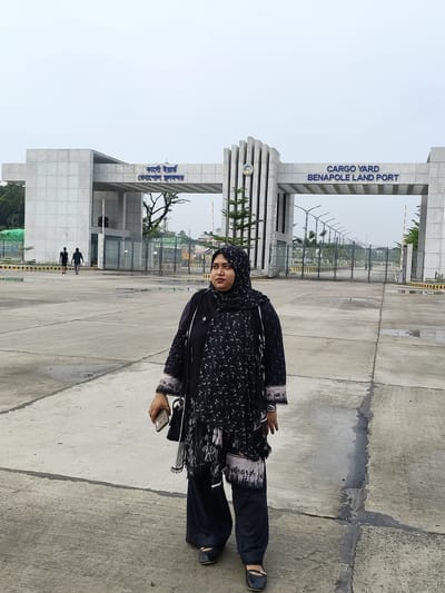 A medium-shot captures a woman standing in front of a large, modern concrete gate structure. The woman is positioned in the right foreground, facing slightly away from the camera and looking to her left. She is wearing a black patterned outfit with a matching headscarf and dark pants. She holds a light-colored mobile phone in her right hand and a dark bag is slung over her left shoulder. Two men, seen from behind, walk away from the camera in the middle ground, heading towards the gate. The gate structure, with text that reads "CARGO YARD BENAPOLE LAND PORT" in English and Bengali, is grand and imposing, featuring a symmetrical design with large concrete pillars and a horizontal beam. The area in front of the gate appears to be a wide, paved courtyard, with some puddles on the ground suggesting recent rain. In the background, beyond the gate, a row of tall streetlights and green trees are visible under a cloudy sky. The overall environment suggests an official entryway to a port facility.
