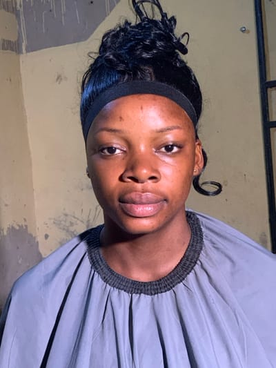 The scene depicts a young adult female, appearing to be of African descent, captured in a medium close-up shot indoors in Garaku, Nigeria. The specific location within Garaku, such as a street or building type, is not discernible from the visual evidence. She is facing directly forward, looking towards the camera. Her hair is styled in a dark, curly updo, secured by a black headband across her forehead. A singular dark curl hangs loosely behind her right ear. She is wearing a grey stylist's cape or similar protective garment, which features a dark, elasticized band around the neckline and visible pleats. The background consists of an interior wall painted in a faded light yellow or beige, showing areas of discoloration, possible paint drips, and plaster irregularities. A dark vertical frame, consistent with a window or door jamb, is visible on the right side of the image. No other individuals or significant objects are present within the frame.