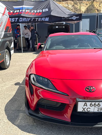 The image shows the front of a red Toyota Supra with the license plate A 666 XC 28 RUS. The car is parked near a tent with the words "FIRST DRIFT TEAM" and "TRAKHAEB Racing." There are people visible under the tent. The location is likely Sadovoe, Russia, based on the license plate information.