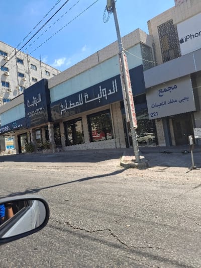The image depicts an urban street scene in Dabouq, Jordan, captured during daylight hours under a clear blue sky with minimal cloud cover.
In the immediate foreground, the left side of a silver vehicle's side mirror is visible, reflecting a partial view of an individual wearing a blue shirt. The asphalt road surface exhibits multiple linear cracks, indicating wear. A long, dark shadow extends across the roadway, originating from the lower right and stretching towards the upper left.
In the midground, a light-grey utility pole stands prominently on the right side of the road, supporting multiple overhead electrical wires. To the left of the pole, a series of commercial establishments line the street. The central storefront features a light blue-grey facade with a darker grey horizontal trim, displaying Arabic signage "الدولية للمطابخ" (International Kitchens) and smaller English text "INTERNATIONAL KITCHENS P.O.". This establishment has large glass display windows. To its immediate right, another storefront bears the Arabic text "مجمع" (Complex) followed by "يحيى مخلد النعيمات" (Yahya Mukhalled Al-Nuaimat) and the numerical address "221". Above this storefront, a partial Apple "iPhone" logo is discernable on the building's upper section. Further to the left, a sign partially reads "STORE".
The background features a multi-story building constructed of light-colored material, likely concrete, with numerous windows and externally mounted air conditioning units visible on its facade. Overhead power lines crisscross the upper portion of the frame against the sky.