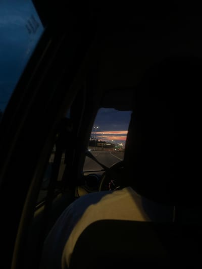 The image depicts a view from the interior of a car, looking out towards a highway at dusk. The sky is a gradient of dark blue and orange, indicating either sunrise or sunset. Streetlights line the highway, and a few cars are visible in the distance. The foreground is dark, with the silhouette of a person's head and shoulder visible on the right side. Part of the car's dashboard and steering wheel are also in view on the left side. The image suggests a journey on a road near Pesochny, Russia, given the context provided.
