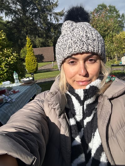 A woman is standing in an outdoor setting, likely a garden or park. She is wearing a gray puffer jacket, a black and white zebra-print fleece sweater, and a black and white knitted beanie with a black pom-pom. Her hair is blonde and she appears to be in her late 20s or early 30s. In the background, there is a small wooden structure and a picnic table set with a green and white checkered tablecloth. There is a bottle of water and food on the table. The setting is in the city of Cloughton, United Kingdom, located at latitude 54.369 and longitude -0.506. The image is a selfie taken from the woman’s perspective. She is looking directly at the camera with a neutral expression. The sun is shining, casting a warm glow on the scene. The woman is likely enjoying a leisurely outdoor experience.