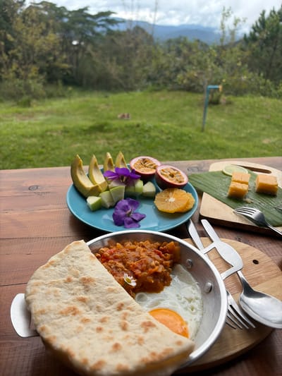 The image depicts a breakfast spread on a wooden table set against a backdrop of lush greenery and rolling hills, suggesting a location in a rural or mountainous area. The foreground features a silver pan containing a fried egg with a visible yolk, a portion of what appears to be a savory stew with diced vegetables, and a piece of flatbread folded over the edge of the pan. To the right, a small wooden cutting board displays three cubed pieces of a yellow, possibly cheese-like food, a lime wedge, and a sprig of something green. Cutlery, including a fork and spoon with a bunny-shaped handle, are also visible.

Further back, a blue plate holds sliced avocado, cubed green fruit (possibly apple or pear), halved passion fruits, sliced orange, and two purple flowers. The environment behind the table is a verdant landscape with trees, grass, and distant mountains under a cloudy sky.

The scene is outdoors, with natural light illuminating the food and the surrounding environment. The overall impression is one of a tranquil, scenic breakfast setting. The specific mention of Ban Khun Wang, Thailand, would imply this natural setting is characteristic of that region, known for its agricultural landscapes and mountainous terrain.