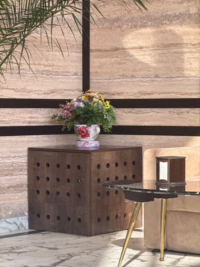 A flower arrangement sits beside a brown cubical table with holes. A coffee table and beige sofa are adjacent. The scene is indoors, on a beige tiled floor with brown stripes. A leafy plant appears in the bottom left corner. A marble-like wall is visible in the upper and right edges. The viewpoint is from directly above.