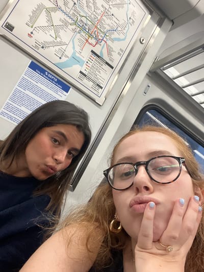 The image depicts two young adult females inside a SEPTA public transit vehicle in Philadelphia, Pennsylvania, United States. Both individuals are centered, looking towards the camera, and posing with pursed lips. The female on the left has long, dark brown hair and is wearing a dark, possibly navy, top. The female on the right has long, reddish-blonde hair, wears tortoise-shell framed glasses, and has a gold-toned ring with a decorative setting on her left ring finger; her fingernails are painted light blue, and her left hand is placed against her chin. In the background, a detailed SEPTA system map of Philadelphia and a "Title VI Notice to the Public" are visible on the vehicle's interior wall. The vehicle's interior features light-colored paneling, overhead lighting, and an overhead luggage rack; a window showing a dark exterior is also partially visible.