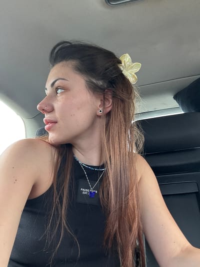 The image features a young woman inside what appears to be a vehicle, likely a car or bus, given the interior paneling and visible light sources. Her head is tilted, and she is looking downwards and slightly to her left, captured in a selfie-like pose.
She has long, light brown or auburn hair, which is pulled back slightly with a translucent, iridescent yellow, flower-shaped hair clip positioned on the top of her head. She wears a small black earring with a silver rim in her left ear, and a silver hoop nose ring. Her complexion is fair, with numerous freckles visible on her face and arm. She has dark, shaped eyebrows and appears to have light-colored eyeliner or eyeshadow on her lower lid. Her lips are slightly parted and appear glossy.
She is wearing a black ribbed tank top. Around her neck are two layered necklaces: one a thin silver chain with a small blue, possibly butterfly-shaped, pendant, and the other a beaded necklace in shades of blue, black, and silver. Attached to the silver chain, beneath the blue pendant, is a small black rectangular tag. Text visible on this tag reads "PAUSE IT'S A S" on the upper line, with "EST." partially visible below it.
The background consists of light gray and dark gray/black surfaces, characteristic of a vehicle's interior. Bright light from what seems to be a window or windshield indicates that it is daytime. Her expression is neutral and appears contemplative. No specific visual cues within the image confirm the location as Slivnitsa, Bulgaria.