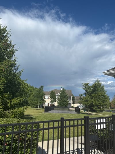 The image shows a residential area with a faint rainbow visible in the sky. In the foreground, there is a black metal fence. Beyond the fence, a manicured lawn is visible, with trees and a two-story building in the background. The building appears to be a residential complex with multiple windows and a light-colored exterior. The sky is a mixture of blue sky and dark clouds, suggesting recent or impending rain. The scene is set during daylight hours.