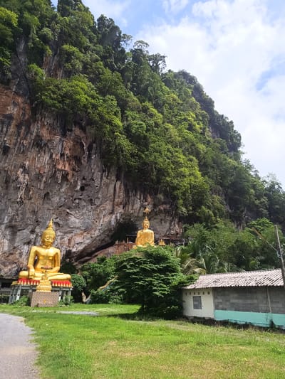 This image shows a landscape in Thap Put, Thailand. Dominating the scene is a large, rocky cliff covered in lush green vegetation. In front of the cliff stand two golden Buddha statues. One statue is on the left, and the other is positioned in the center. To the right of the statues is a small, single-story building constructed of gray cinder blocks and partially painted white. The building has a roof made of tiles. The foreground is filled with green grass and a gravel path. The sky above is partly cloudy.