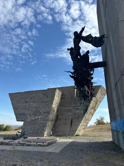 The image depicts the "Monument to the Liberators of Izium" located on Mount Kremenets in Izium, Kharkiv Oblast, Ukraine. The scene is dominated by brutalist concrete structures and bronze sculptures under a clear blue sky with scattered white cumulus clouds.
In the center-right, a large, dark bronze sculpture protrudes from a vertical concrete wall. This complex sculpture features numerous interwoven figures, appearing to be soldiers or combatants, some with weapons such as rifles or spears. The uppermost figure stands with an arm raised, accompanied by a tattered flag-like element. Other figures are depicted in dynamic, entangled poses below. The concrete wall it is affixed to is light gray and has a horizontal blue stripe near its base on the far right edge of the frame.
To the left, a large, angular concrete wall with visible formwork seams forms part of the monument. Graffiti in black spray paint is present on its lower section. In front of this wall, on a low, damaged concrete platform with exposed rebar, lies a smaller bronze sculpture of a recumbent figure, possibly a fallen soldier, accompanied by an object that resembles a helmet or weapon.
The ground consists of a paved area with square concrete pavers and rough cobblestones. In the background, beyond the concrete structures, is a landscape of green hills and vegetation under the bright sky. A faint white contrail is visible in the sky.
