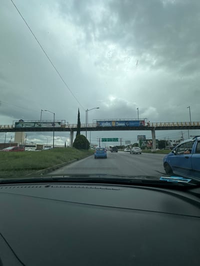 The image depicts a multi-lane urban highway in San Luis Potosí City, Mexico, specifically on Avenida Salvador Nava Martínez (Periférico), looking southeast towards its intersection with Highway 57 (Carretera Central), where routes to Matehuala and Guadalajara are indicated.
The scene is captured from the interior of a vehicle, evidenced by the dark dashboard and windshield in the foreground. The windshield exhibits water droplets, suggesting recent rain or high humidity. The sky is completely overcast with thick, grey clouds. A power line traverses the upper portion of the frame.
The highway is a wide, asphalt road with at least three lanes of traffic visible, heading away from the viewer. The road surface appears damp. To the immediate right, a light blue hatchback or small SUV is partially visible, either parked on the shoulder or stopped in the rightmost lane, with its front passenger wheel turned. Further ahead in the middle lane, a blue hatchback is visible, driving forward. Several other vehicles of various colors are distributed in the distant lanes.
A large, grey pedestrian overpass with yellow railings spans the highway. Two large billboards are mounted on its structure: the left billboard displays a green background with a "MONTESSORI" logo, while the right billboard features the word "FORMA" in large, dark blue letters on a lighter blue and white background. A single individual is visible walking on the pedestrian bridge, moving from right to left across the far side of the right billboard.
In the grassy median dividing the highway, a tall, slender, conical conifer tree stands centrally, with a smaller, rounded dark green bush at its base. Overhead, a large green gantry sign with two directional arrows is visible in the distance; the left arrow points to "SLP / Matehuala," and the right arrow points to "SLP / Guadalajara." Further down the road on the right, another billboard with a green background partially reads "SALUD" and "AMOR." On the left side of the road, beyond the median, a large, light-colored building is partially visible with red vehicles parked nearby. A blue or teal-colored paper object is visible tucked under the passenger side windshield wiper blade.