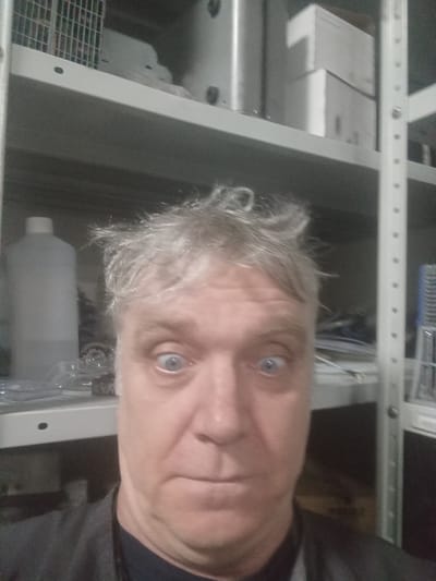 A medium shot photograph captures a Caucasian man in his 50s or 60s with messy, graying hair and wide, blue eyes. He is looking directly at the camera, with his mouth slightly open in an expression that could be interpreted as surprised or taken aback. The man is wearing a dark gray jacket over a navy blue t-shirt.
The background suggests an indoor storage or workshop area. Visible are several shelves filled with various items. On the shelf closest to the man, a large, translucent white bottle with a white cap is positioned to his left. To his right, a metal shelf support is visible, with items on shelves above and below that are partially obscured. The shelves themselves appear to be made of light-colored metal, common in industrial or storage settings. The overall lighting is dim, with the man's face being the primary illuminated subject. The image does not contain information about the city of Nikopol, Ukraine.