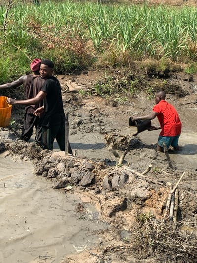 Two men collect water near a muddy riverbank. The first man, in a red shirt, pours water from a black container. The second man, wearing a black shirt and a red cap, stands near an orange bucket. Thick vegetation borders one side of the riverbank. The ground is muddy, interspersed with dry branches. The river itself is a light gray color.