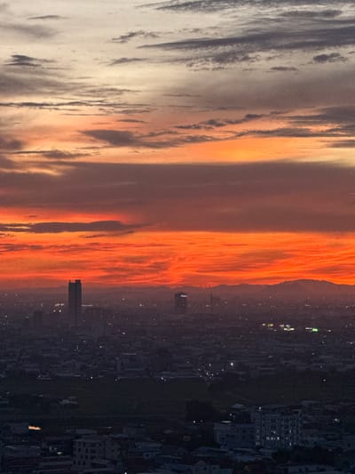 This is a vertical, wide-angle photograph of a city skyline at dusk, with a dramatic sunset filling the sky. The sun has set, casting a vibrant orange and yellow glow on the clouds. The clouds are layered, with some appearing wispy and light, while others are thicker and more defined. The cityscape below is dark and appears to be a densely populated urban area with many buildings, some of which are illuminated by interior lights. There is a prominent skyscraper visible in the mid-ground. The scene appears to be captured from a high vantage point, looking down and across the city. The time of day is clearly dusk, as evidenced by the sunset colors and the illuminated city lights. The weather appears to be clear, allowing for a vibrant sunset. The location is identified as Khan Mean Chey, Cambodia, which is a district within Phnom Penh. The image evokes a sense of urban grandeur and the beauty of nature juxtaposed. There is no visible text in the image.