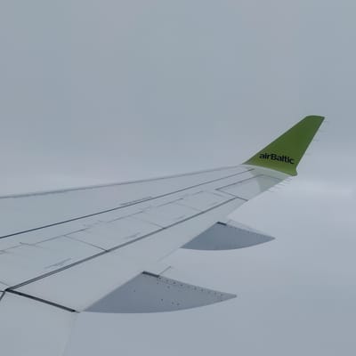 The image displays the right wing of an airBaltic aircraft in flight, observed from a passenger window. The wing is predominantly white, featuring multiple visible control surfaces, panels, and structural lines across its upper surface. A bright green winglet, curving upwards from the wingtip, prominently displays the "airBaltic" logo in black letters. Several small, thin, pointed protrusions, likely static dischargers, are visible along the trailing edge of the green winglet. The background is a uniform, light grey sky, indicating overcast conditions or flight through clouds/haze. No ground features, other aircraft, or specific landmarks are visible, suggesting the aircraft is at cruising altitude. The lighting is soft and diffused, with minimal distinct shadows on the wing surfaces. Faint, dark textual markings are present on the white wing panels, but their content is illegible. The image does not provide visual cues to confirm the aircraft's location over Ķesterciems, Latvia.