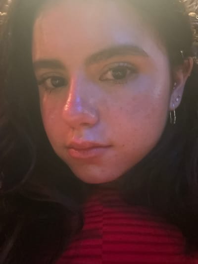 A close-up, rotated portrait features a young person, likely female, looking directly at the camera with a neutral expression. The subject's head is tilted significantly, with their dark hair framing the face. Highlights are visible on the forehead and bridge of the nose, suggesting direct, possibly artificial, light. Several ear piercings, including multiple studs and a small hoop-style earring, are visible on the upper ear. A red garment with vertical stripes, possibly a top or sweater, is visible on the right side of the frame. The background is dark and indistinct, indicating an indoor setting. No specific location cues, weather conditions, or text are discernible.