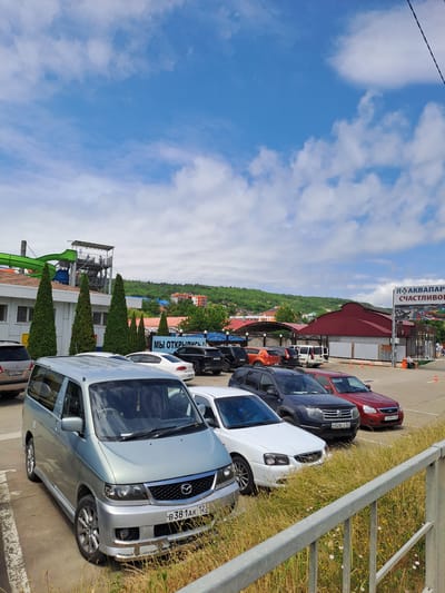 The image shows a parking lot outside an aquapark in Dzhubga, Russia. The sky is blue with scattered white clouds. In the foreground, there is a silver minivan and several other cars parked. Behind the cars, there is a building with a water slide extending from its roof. A sign at the entrance to the aquapark reads "АКВАПАРК СЧАСТЛИВОЙ" (Aquapark Happy). The scene appears to be a sunny day, likely during the summer season when the aquapark would be open. Green trees and hills are visible in the background, suggesting a coastal or resort town setting. There are no people clearly visible in the immediate foreground, but a few figures can be seen in the distance near the aquapark entrance.