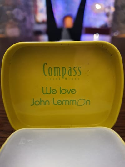 An open, yellow tin with "Compass Fresh Mints" and "We love John Lemmon" embossed in green text is shown. The tin is placed on a wooden surface. The background is blurred and shows a dimly lit interior with colorful lights. The city of Malia Municipal Unit, Greece is not visible in the image.