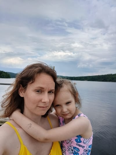 A woman and a young child are taking a selfie on a wooden pier. The woman, with brown hair, wears a yellow top. The child, also with brown hair, wears a pink swimsuit with blue characters. They are embraced, with the child's arms around the woman's neck. The background shows a large body of water, a forested shoreline, and a cloudy sky. The location is the city of Revda, Russia.