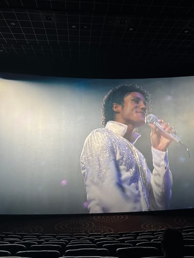 The image shows a close-up of a person singing into a microphone. The person has dark, curly hair and is wearing a white jacket with sparkly embellishments. The person's head is tilted upwards and they are looking towards the microphone. The background appears to be a stage setting with bright lighting. The image is cropped on the left and right edges showing the room. The location is not possible to determine based on this image.
