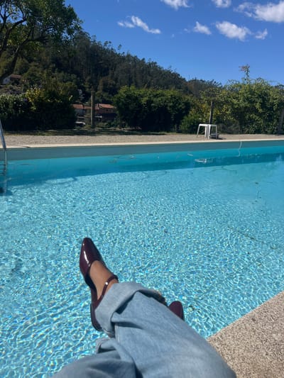 A low-angle shot captures the lower body of an individual, seen from the waist down, seated by a swimming pool in Oliveira de Frades, Portugal. The subject's legs are extended forward, with feet submerged in the clear, rippling blue water of the pool. The person wears light blue denim jeans, cuffed at the ankles, and dark red, pointed-toe flats with ankle straps. A thin, dark bracelet is visible on the left ankle.

The foreground is dominated by the swimming pool, featuring a light-colored, speckled concrete coping. A stainless steel ladder rail is partially visible on the left side of the pool. A white cable or hose floats on the surface of the water near the far edge. Beyond the pool, a narrow strip of light-colored ground separates it from a dense line of green foliage, including shrubs and trees. Further in the background, a steep, heavily forested hillside rises, covered with dark green trees. Scattered among the trees and on the hillside, several residential structures with reddish-brown roofs are visible. The sky is bright blue with scattered white cumulus clouds. On the ground behind the pool, towards the right, a white plastic stool is positioned near a thin, upright pole.