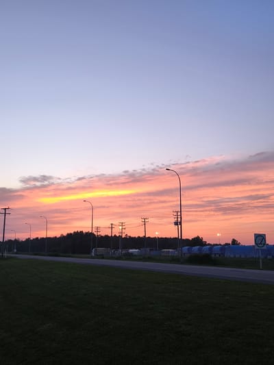 The image depicts a rural or industrial area in Mirabel, Canada, during a sunset. The sky displays a gradient of soft blues and purples, transitioning into vibrant oranges and yellows in the lower portion, with thin, wispy clouds illuminated by the setting sun. In the foreground, a dark green, well-maintained grassy area slopes down towards a paved road. The road runs horizontally across the frame, with a tree line of dark green foliage forming a backdrop.
Several utility poles with electrical wires are visible along the roadside. Streetlights are also present, some illuminated. On the other side of the road, there are industrial or storage structures covered in blue tarps, some appearing to be temporary or modular buildings. A few trucks and a white utility vehicle are parked near these structures. A regulatory sign with a green circle and a white pictogram of a person in a seatbelt is posted on a pole on the right side of the road. The overall scene suggests an outdoor setting, likely on the outskirts of a town or in an industrial park, given the presence of infrastructure and storage facilities, with the natural beauty of the sunset as a prominent feature. No people are visibly present in the image.