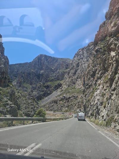 The image depicts a road winding through a mountainous landscape in Frati, Greece. A white van is visible driving on the asphalt road. The road is bordered by a metal guardrail on one side and sheer rock cliffs on the other. Vegetation clings to the rock faces. The sky is blue with reflections visible on a window.
