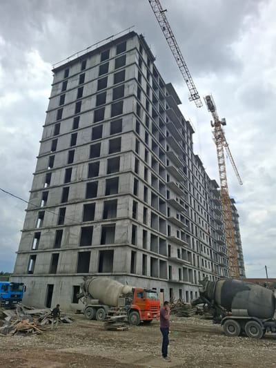 An unfinished concrete apartment building, appearing to be in the city of Argun, Russia, dominates the frame under a cloudy sky. Two large construction cranes, one yellow and the other red, are visible near the building. In the foreground, an orange concrete mixer truck is parked, and to its left, a blue truck is also present. A pile of lumber and debris is scattered on the ground. A single man in a dark shirt and pants stands near the concrete mixer, seemingly on his phone. Another individual, wearing a dark shirt and cap, is standing near the blue truck. The ground is covered in gravel and dirt, indicating an active construction site.