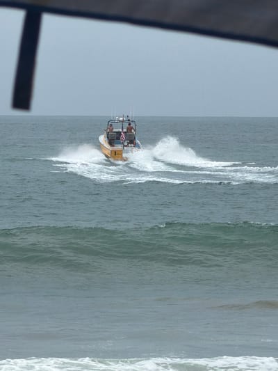 An offshore shot captures a yellow and white boat with two individuals on board, heading away from the viewer. The boat is leaving a white spray wake behind it, indicating movement through the water. The ocean appears choppy with moderate waves, and the sky is overcast and hazy, suggesting cloudy weather. The foreground of the image is dominated by the ocean, with the horizon line barely visible in the distance, separating the sea from the pale sky. The presence of a dark, angled object in the upper left corner of the frame suggests the photo was taken from an elevated vantage point, possibly from a structure or another vessel. The overall scene depicts a marine environment with a boat actively navigating the sea.