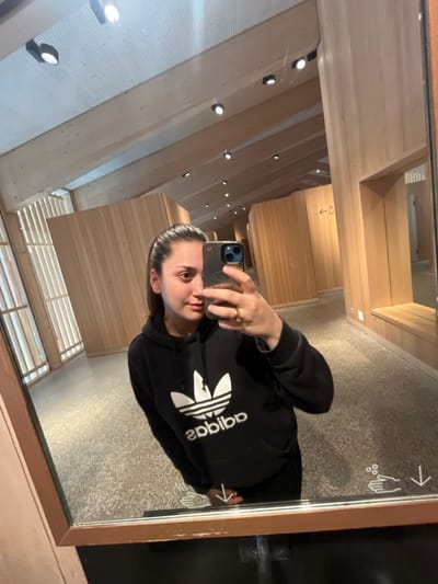 An image taken as a mirror selfie depicts a young adult female within a modern public facility in Erstfeld, Switzerland, likely a restroom or changing area. The subject, positioned centrally, is reflected from the chest up, looking directly towards the camera held in her left hand. She has dark hair pulled back from her face, a neutral expression, and is wearing a black hoodie with a prominent white Adidas Trefoil logo. A gold-colored ring is visible on her left ring finger.
The background, seen through the mirror, reveals an interior constructed predominantly with light-colored, vertical wooden paneling on the walls and a slatted wooden ceiling with multiple recessed spotlights. The floor is a dark, textured material, possibly polished concrete or terrazzo. To the subject's right, a black arrow points left towards a universal male/female restroom symbol on a wooden wall. Below the mirror, on the surface it rests upon, white pictograms indicate handwashing functions: two hands under a downward arrow, and another showing hands with bubbles and a downward arrow, suggesting the location of water and soap dispensers, respectively. The mirror itself has a light-colored wooden frame.