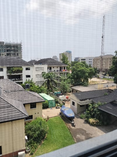 This image presents an elevated view of a mixed residential and urban landscape in Falomo, Nigeria, under a bright, hazy sky.

In the immediate foreground, a blurred window frame or ledge is visible. The scene below features several buildings with dark-tiled, pitched roofs. To the lower left, a light-colored two-story building is situated next to a well-maintained green lawn. External air conditioning units are affixed to its wall. Adjacent to this, a paved area extends, containing a blue tarp-covered boat resting on a trailer. Another light-colored building with a dark-tiled roof is present to the right of the paved area, surrounded by sparse greenery.

In the mid-ground, a cluster of multi-story residential buildings, primarily white or light-colored, features balconies. One balcony displays several potted plants, while another has clothes draped over a railing. Black water tanks are visible on some rooftops. Behind these residences, an open area contains at least two green shipping containers, various tanks, and other industrial equipment.

Further in the background, to the far left, a multi-story building under construction is partially visible, encased in scaffolding. Towards the center-right background, a prominent tall, lattice-style communication tower stands. Surrounding the tower are additional multi-story buildings, some appearing residential and others more modern high-rise structures. An unpaved, cleared lot is also present. Numerous trees and lush vegetation are dispersed throughout the urban environment. No individuals or specific actions are discernible.