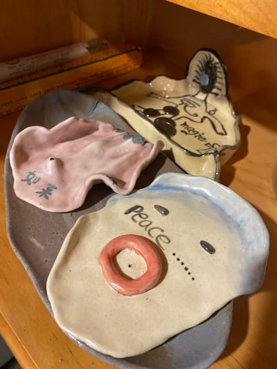 A close-up, overhead shot captures three small, decorative ceramic dishes on a wooden surface. The dishes appear to be handmade, with irregular shapes and surfaces.

The largest dish, in the foreground and slightly to the right, is light tan with a light blue rim. It has two dark oval indentations that resemble eyes and a string of small black dots below them, suggesting a face. In the center of the dish is a raised, circular object with a red glaze, resembling a mouth or a ring. The word "Peace" is written in black script across the surface of this dish.

Behind and to the left of the "Peace" dish is another ceramic piece, this one light tan with a wavy, irregular edge. It features a drawing of a figure with wings and a circular emblem. The word "magie" is written in black script on this piece, along with a stylized symbol resembling a cross with a circle.

The smallest dish, positioned in the center and front, is a delicate pink with a ruffled edge. It has a small raised point in the center and appears to be designed to hold something. Black characters, possibly Chinese, are inscribed on its surface.

The overall impression is one of whimsical, handcrafted art pieces. The lighting suggests an indoor setting, possibly a shelf or table, with a warm wooden background. There are no indications of weather or a specific time of day. The setting is implied to be Dujiangyan City, China, although no specific landmarks or cultural cues are visible in the image itself to confirm this.