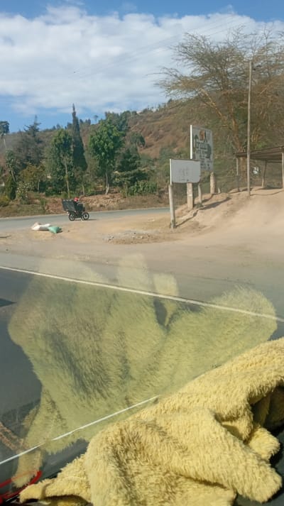 A road scene features a motorbike with a large box mounted on the back driving along the asphalt. Sacks are placed on the roadside. A signboard stands near the intersection, flanked by a sun shelter structure. A hillside with trees rises in the background under a blue sky with scattered clouds. The foreground is dominated by a fuzzy yellow cloth draped over what seems to be the dashboard of a car.