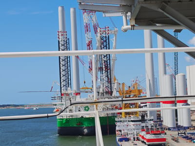 This image captures a bustling industrial scene at a port, likely in Esbjerg, Denmark, given the context. The foreground is dominated by the railing of a vessel, with a clear blue sky overhead and a calm, dark blue sea below.
The main subjects are several large, white cylindrical structures, likely components of wind turbines, standing tall against the sky. Behind them, a complex array of industrial machinery is visible, including massive cranes with red and white and yellow detailing. A prominent green and black ship, named "NORSE WIND", is docked, suggesting ongoing construction or maintenance of wind energy infrastructure. Another white ship, with red and white detailing on its deck, is also present.
The scene depicts a hive of industrial activity, with the presence of cranes and large components indicating the assembly or transportation of wind turbine parts. The bright sunlight and clear blue sky suggest it is a fair weather day, likely in the daytime. The overall atmosphere is one of productivity and large-scale engineering.
Visible text includes "NORSE WIND" on the green ship and "SAFETY STARTS WITH YOU AND ME" on the white ship. The word "VESTAS" is also visible on one of the white cylindrical components. A distant windmill is visible on the horizon, reinforcing the wind energy theme.