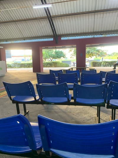 The image depicts an outdoor, covered common area, characteristic of a university campus or large public institution, situated in Lauro De Freitas, Brazil. The area features a light-colored corrugated metal roof supported by dark metal trusses and a fluorescent lighting fixture. The floor is composed of a light-toned, possibly concrete, paved surface. Structural elements consist of dark reddish-brown pillars and beams that create large, open framed portals leading to an exterior environment.
The primary subjects are multiple rows of fixed-mount seating. These are dark blue plastic chairs with black metal frames, typically configured in units of three or more seats. The chairs in the foreground and midground are unoccupied. In the background, through the open portals, a landscaped area with green trees, bushes, and paved sections is visible. Strong natural sunlight enters from the left, creating prominent shadows across the floor and illuminating the exterior.
One individual is partially visible on the far right, seated on one of the blue chairs, facing away from the viewer. Further in the background, a long, dark table with associated benches is discernible, with what appears to be another person or small group nearby, though details are obscured by distance and lighting. On the far left, a segment of a light-colored vertical panel or sign with a partial green letter "N" is visible. The overall scene suggests a quiet moment within a public or educational setting, such as a university campus like UNIME - União Metropolitana de Educação e Cultura, which is present in Lauro de Freitas.