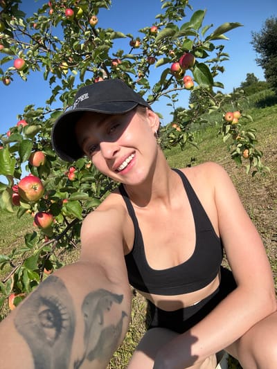In a sun-drenched orchard, a young woman is captured in a selfie, surrounded by the bounty of an apple tree. She wears a black sports bra and a black baseball cap, tilted to partially obscure her face. Her smile is bright, revealing a row of white teeth, and her eyes, one slightly more visible than the other, are focused towards the camera. A large tattoo of an eye is prominent on her forearm, adding a personal detail to the scene.
The apple tree is laden with ripe apples, their red and green hues contrasting with the lush green leaves and the clear blue sky above. Sunlight filters through the branches, dappling the ground with light and shadow. The setting appears to be a rural landscape, with a hint of grass and trees visible in the background, suggesting a warm, pleasant day. The orientation of the image, tilted to the side, gives it a candid and energetic feel. The image conveys a sense of joy and engagement with nature, possibly during a leisurely day out, perhaps apple picking. The text "ZONE" is visible on the side of her baseball cap.
