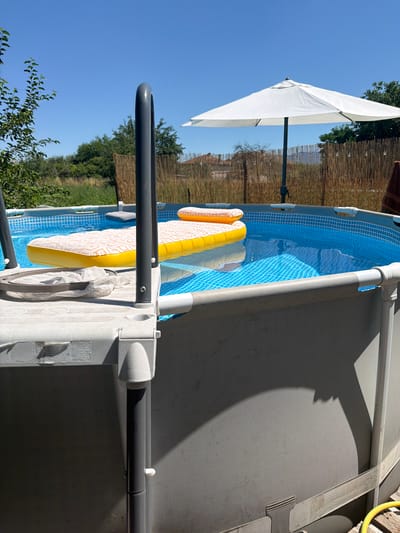 This image depicts an above-ground swimming pool on a sunny day in Karadjovo, Bulgaria. The pool is filled with clear blue water and has a patterned yellow and white inflatable mattress floating on the surface. To the left of the pool, a grey ladder with curved handrails is visible, with a white net attached to the top step. A white patio umbrella stands open behind the pool, providing shade. The surrounding environment features a bamboo fence, green trees, and residential buildings in the distance. The scene is characteristic of a private backyard setting during warm weather.